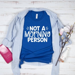 Pu&ograve; includere: Una maglietta blu reale con testo bianco che recita "Not a Morning Person". La maglietta &egrave; piegata e appoggiata su una superficie di legno bianco con una camicia a righe blu e bianche, un portafoglio rosa, un ramoscello di fiori bianchi e una bottiglia di vetro trasparente.