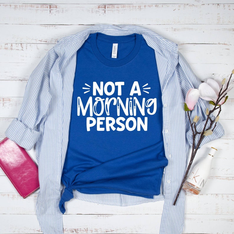 Pu&ograve; includere: Una maglietta blu reale con testo bianco che recita "Not a Morning Person". La maglietta &egrave; piegata e appoggiata su una superficie di legno bianco con una camicia a righe blu e bianche, un portafoglio rosa, un ramoscello di fiori bianchi e una bottiglia di vetro trasparente.