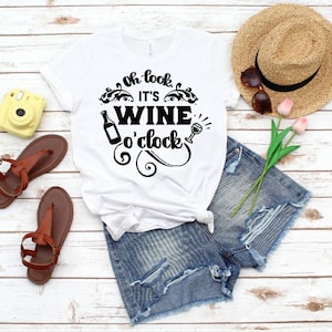 May include: White t-shirt with the text "Oh look it's WINE o'clock" in black lettering. The shirt is on a wooden surface with a pair of brown sandals, a yellow camera, a straw hat, and a bouquet of pink flowers.