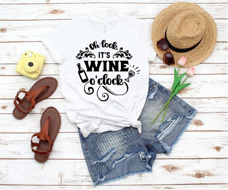 May include: White t-shirt with the text "Oh look it's WINE o'clock" in black lettering. The shirt is on a wooden surface with a pair of brown sandals, a yellow camera, a straw hat, and a bouquet of pink flowers.