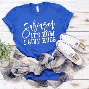 Puede incluir: Una camiseta azul real con el texto "Sarcasm It's How I Give Hugs" en letras blancas.