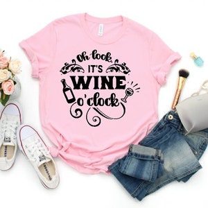 May include: Pink t-shirt with black text that reads "Oh look it's wine o'clock". The text is surrounded by a decorative design. The shirt is laid out on a white surface with a pair of white sneakers, a pair of blue jeans, a makeup brush, and a small gray pouch.
