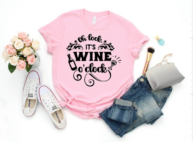 May include: Pink t-shirt with black text that reads "Oh look it's wine o'clock". The text is surrounded by a decorative design. The shirt is laid out on a white surface with a pair of white sneakers, a pair of blue jeans, a makeup brush, and a small gray pouch.