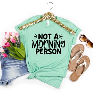 Pu&ograve; includere: Una maglietta verde menta con il testo "NOT A MORNING PERSON" in lettere nere.