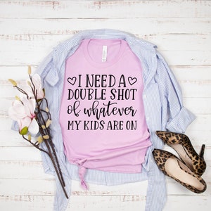May include: A pink t-shirt with the text "I need a double shot of whatever my kids are on" printed on it. The shirt is folded and laid on a white wooden surface with a blue and white striped shirt and leopard print shoes.