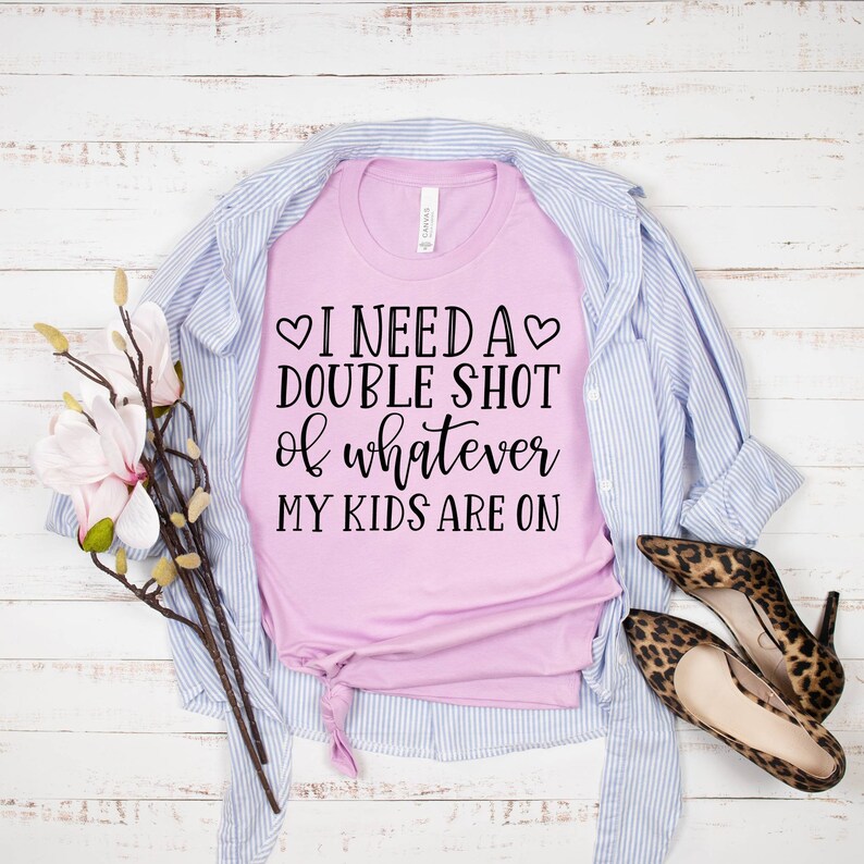 May include: A pink t-shirt with the text "I need a double shot of whatever my kids are on" printed on it. The shirt is folded and laid on a white wooden surface with a blue and white striped shirt and leopard print shoes.