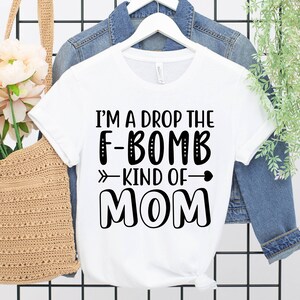 May include: White t-shirt with black text that reads "I'm a drop the F-bomb kind of mom" with an arrow pointing to the right and a heart.