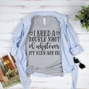 May include: A gray t-shirt with the text "I need a double shot of whatever my kids are on" printed on it. The shirt is folded and laid on a white wooden surface with a blue and white striped shirt, sunglasses, and a potted plant.