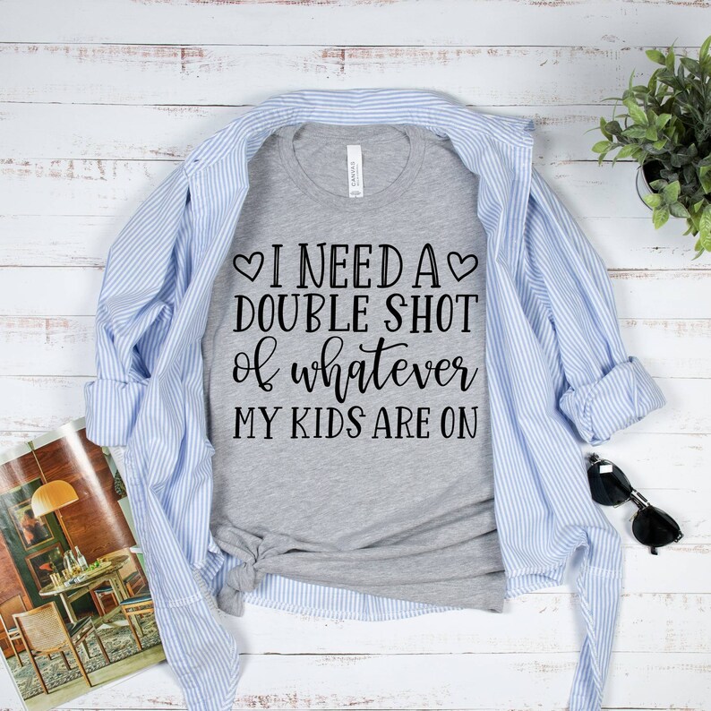 May include: A gray t-shirt with the text "I need a double shot of whatever my kids are on" printed on it. The shirt is folded and laid on a white wooden surface with a blue and white striped shirt, sunglasses, and a potted plant.