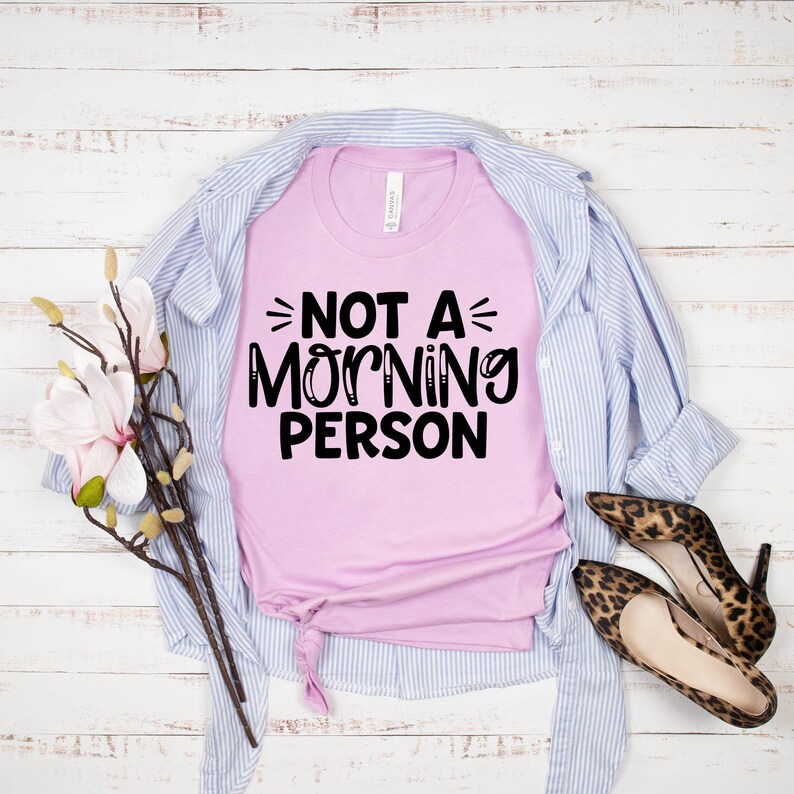 Pu&ograve; includere: Una maglietta rosa con il testo "Not a Morning Person" in lettere nere. La maglietta &egrave; appoggiata su una superficie di legno bianco con una camicia a righe blu e bianche e un paio di scarpe leopardate.