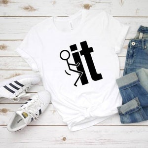 May include: White t-shirt with a black stick figure flipping the bird and the word "it" printed on it. The shirt is laid out on a white wooden surface with a pair of white sneakers and blue jeans.