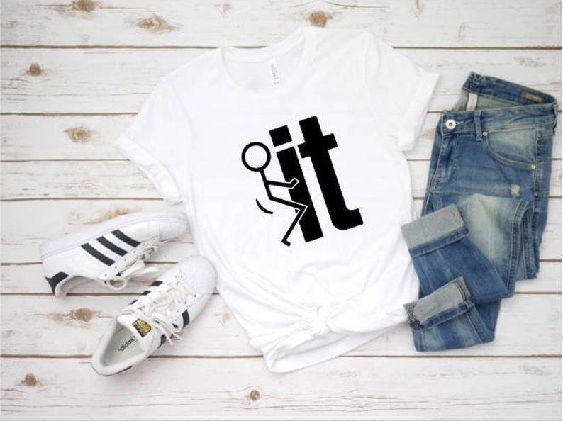 May include: White t-shirt with a black stick figure flipping the bird and the word "it" printed on it. The shirt is laid out on a white wooden surface with a pair of white sneakers and blue jeans.