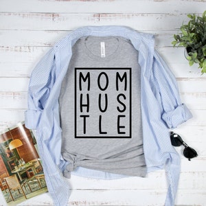 May include: A grey t-shirt with the text "MOM HUSTLE" printed in black within a black square.