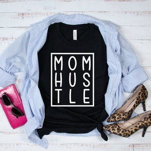 May include: Black t-shirt with white text that reads "MOM HUSTLE" in a box. The shirt is laid out on a white wood surface with a blue and white striped shirt, a pink wallet, and leopard print shoes.