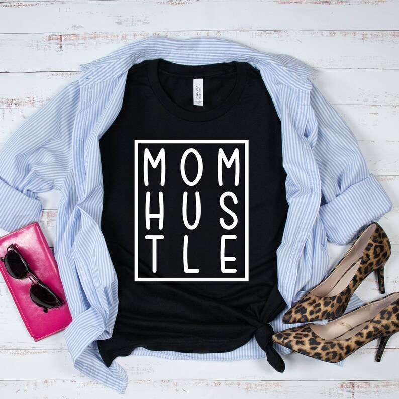 May include: Black t-shirt with white text that reads "MOM HUSTLE" in a box. The shirt is laid out on a white wood surface with a blue and white striped shirt, a pink wallet, and leopard print shoes.
