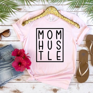 May include: Pink t-shirt with a black square graphic that says "MOM HUSTLE" in block letters.