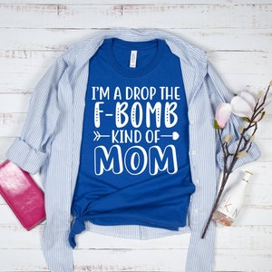 May include: A royal blue t-shirt with white text that reads "I'm a drop the F-bomb kind of mom" with a white arrow pointing to the right and a white heart.
