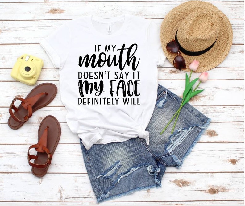 May include: White t-shirt with black text that reads "If my mouth doesn't say it my face definitely will". The shirt is lying on a white wooden surface with a pair of brown sandals, a yellow camera, a straw hat, and a pair of denim shorts.