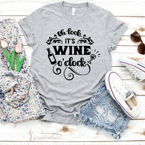 May include: Gray t-shirt with the text "Oh look, it's wine o'clock" in black. The text is surrounded by a decorative design with a wine bottle and a wine glass.