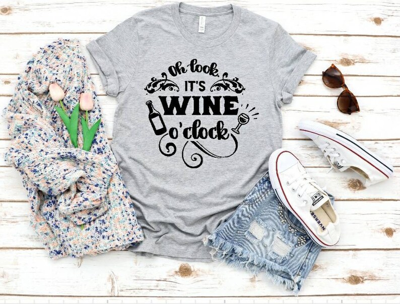 May include: Gray t-shirt with the text "Oh look, it's wine o'clock" in black. The text is surrounded by a decorative design with a wine bottle and a wine glass.