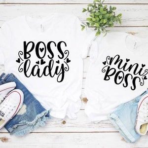 May include: Two white t-shirts with black text reading "BOSS lady" and "mini BOSS". The shirts are styled with denim shorts and white trainers. The design of the shirts is casual and playful.