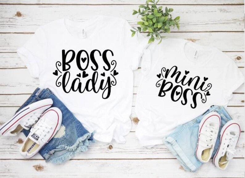 May include: Two white t-shirts with black text reading "BOSS lady" and "mini BOSS". The shirts are styled with denim shorts and white trainers. The design of the shirts is casual and playful.