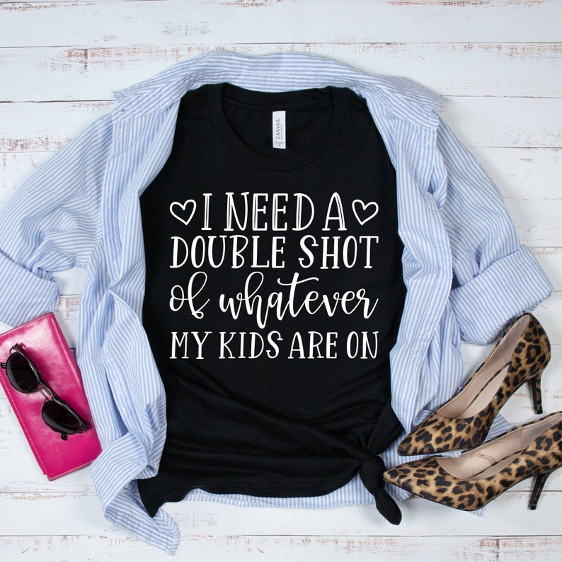 May include: Black t-shirt with white text that reads "I need a double shot of whatever my kids are on". The shirt is folded and laid on a white wood surface with a blue and white striped shirt, a pink wallet, and a pair of leopard print shoes.