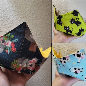 May include: Three fabric bowl cozies with yellow trim. The cozies are made of cotton fabric with different patterns. One features a koala design, another has black cats, and the third has black and white cows.