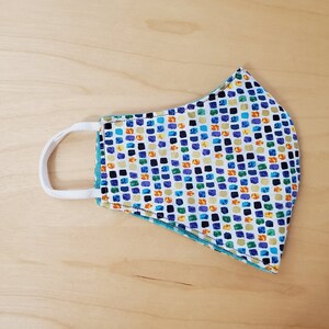 May include: A white face mask with a colorful geometric pattern of squares in shades of blue, green, yellow, and orange. The mask has a white elastic band.