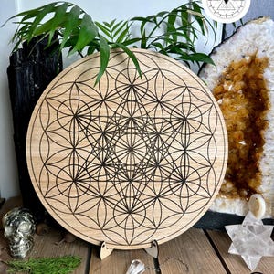 May include: A circular wooden board with a geometric pattern, supported by a wooden stand. The board has a detailed black design on a light wood surface. The text "TASMAN OAK" is visible. The image also includes crystals and plants.