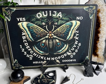 May include: A black wooden Ouija board with a green and gold butterfly design. The board features the letters of the alphabet, numbers 1-9 and 0, and the words "YES", "NO", "HELLO", and "GOODBYE".