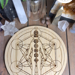 May include: A round, light-colored bamboo board with an engraved human figure and geometric patterns. The figure has seven chakra symbols aligned along the body. The text "10MM BAMBOO" is at the bottom. The board is surrounded by crystals and plants.