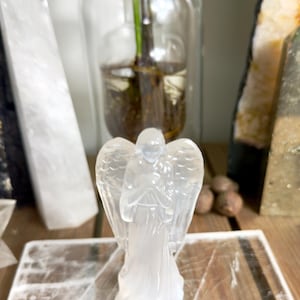 May include: A translucent angel figurine with outstretched wings. The angel is standing on a light-coloured, textured surface. The background includes a glass vase with green plants and other decorative items.