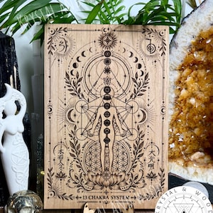 May include: A rectangular wooden plaque featuring a laser-engraved 13 chakra system diagram. The design includes a central human figure, geometric patterns, moon phases, and floral motifs. The text 13 CHAKRA SYSTEM is printed at the bottom.