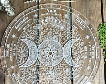 May include: A clear, circular decorative piece with intricate white etched designs. The design includes phases of the moon, floral patterns, and text indicating seasonal celebrations like Samhain, Yule, and Ostara. The piece is on a wooden surface.