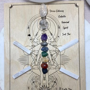 May include: Wooden board with a chakra layout, featuring a series of colorful gemstones and clear quartz crystals. The design includes geometric patterns and the text "Divine Gateway" and "Earth Star". White selenite wands are placed around the central design.