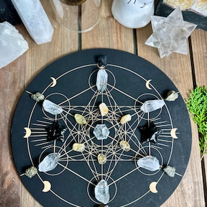 May include: A black Metatron's Cube grid with gold geometric lines, crescent moons, and various crystals. Crystals include clear quartz, black tourmaline, and pyrite. The grid is on a wooden surface.