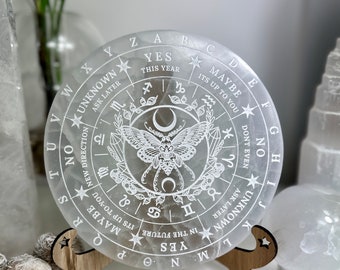 May include: White selenite crystal pendulum board with a moth design and zodiac symbols. The board is on a wooden stand. The text on the board reads "Yes, This Year, Maybe, Its Up To You, New Direction, Unknown, Ask Later, Don't Even, No, Unknown, Ask Later".