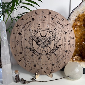 Könnte beinhalten: Ein rundes Holz-Divinationsbrett mit einem schwarz-weißen Mottiv. Das Brett hat ein rundes Zifferblatt mit Buchstaben und Wörtern wie "Ja", "Nein", "Vielleicht", "Später fragen" und "Unbekannt".