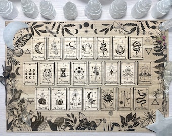 May include: A wooden board with a black and white illustration of the Major Arcana tarot cards. The cards are arranged in a grid pattern and feature various symbols and imagery, including the Fool, the Magician, the Empress, the Emperor, the Hierophant, the Lovers, the Chariot, Strength, the Hermit, Wheel of Fortune, Justice, the Hanged Man, Death, Temperance, the Devil, the Tower, the Star, the Moon, the Sun, Judgement, and the World.