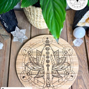 May include: A round, wooden decorative piece with a detailed engraved design featuring a human figure within a lotus flower, surrounded by geometric shapes and chakra symbols. The text "THICK TAS OAK" is at the bottom. The piece is on a wooden surface.