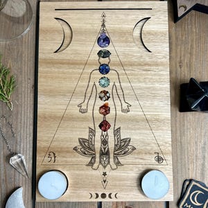 Op de afbeelding: Houten chakra bord met een lasergeëtste menselijke figuur, versierd met kleurrijke edelsteen representaties van de chakra's. Het bord heeft halve maan uitsparingen, een lotusbloem en symbolen. Twee waxinelichtjes zijn onderaan geplaatst.