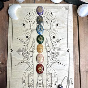 Könnte beinhalten: Ein Holz-Chakra-Brett mit einer detaillierten Illustration einer menschlichen Figur und sieben farbigen Steinen, die entlang des Körpers ausgerichtet sind. Ein lila Pendel hängt an der Seite. Zusätzliche Kristalle und ein mondförmiger Stein sind ebenfalls vorhanden.