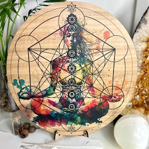May include: A round wooden mandala with a colorful watercolor design of a person in a meditative pose. The design includes geometric shapes and symbols, including a flower of life pattern.