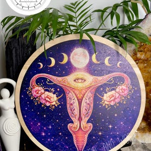 May include: Round wooden wall art featuring a celestial design with a uterus illustration. The artwork has a dark blue background with a galaxy pattern, a full moon, crescent moons, and floral accents. The text "Krystal & Wood" is visible.