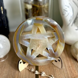 以下が含まれることがあります： 星型の切り抜きが施された、彫刻されたアゲートクリスタル球体。木製のスタンドに展示されています。クリスタルは白、黄色、茶色の帯模様です。乾燥したラベンダーとクリスタルペンデュラムが前景にあります。