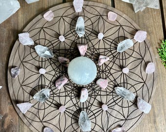 May include: A wooden crystal grid with a geometric design, featuring a central sphere and various crystals in shades of pink, clear, and white. The grid is set on a wooden surface, with additional crystals and objects in the background.