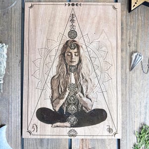May include: A wood-burned mandala design with a woman meditating in the center. The mandala features a triangle with a circle at the top and seven chakras with their corresponding Sanskrit symbols. The text "本質" is written at the top of the triangle.