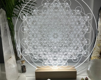 May include: A white geometric design, known as the Flower of Life, is illuminated on a clear acrylic disc. The disc is mounted on a wooden base. The design is surrounded by crystals and other decorative items.