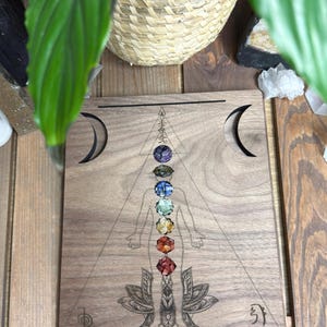 May include: A rectangular wooden board featuring a laser-engraved human silhouette centered within a triangle. Seven small, multi-colored gemstone chips are arranged vertically along the torso. Two circular white tea light candles sit at the bottom.